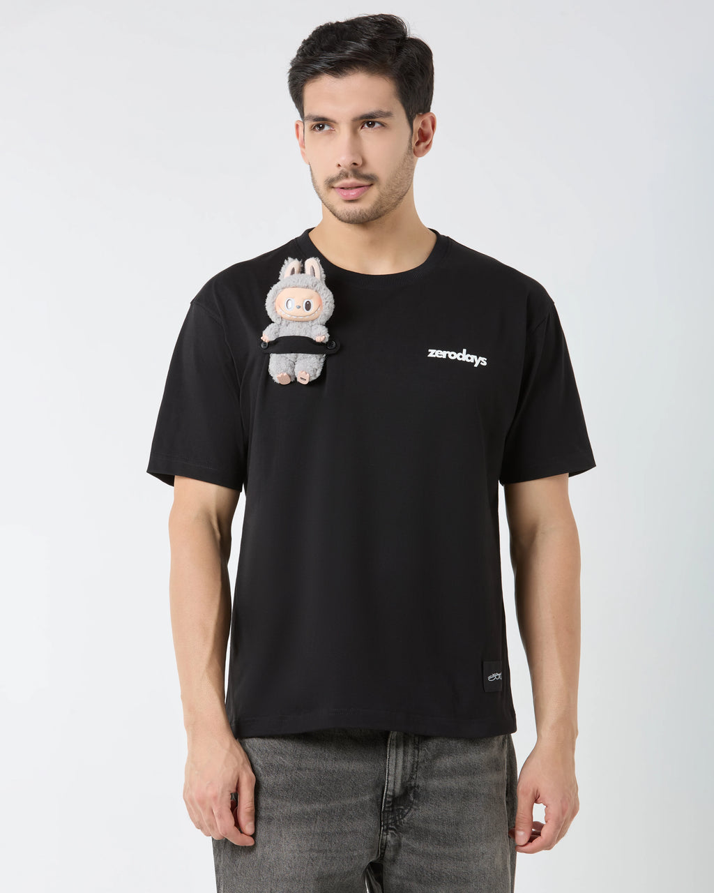 Man wearing a black t-shirt with a small plush toy attached, on a plain background
