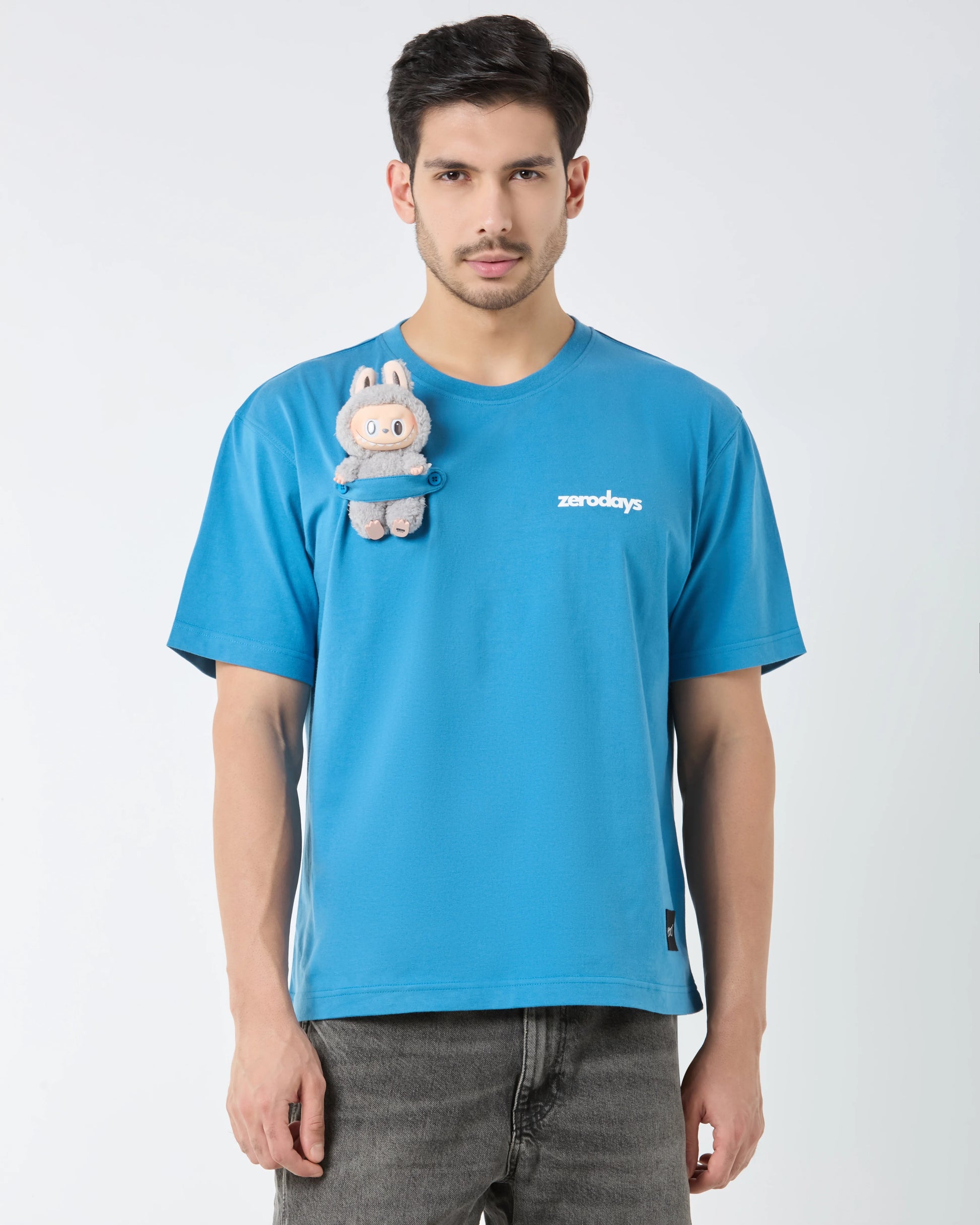 Man wearing a blue t-shirt with a small plush toy attached, on a plain background
