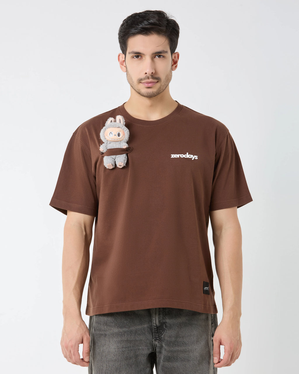 Man wearing a brown t-shirt with a small plush toy attached, on a plain background