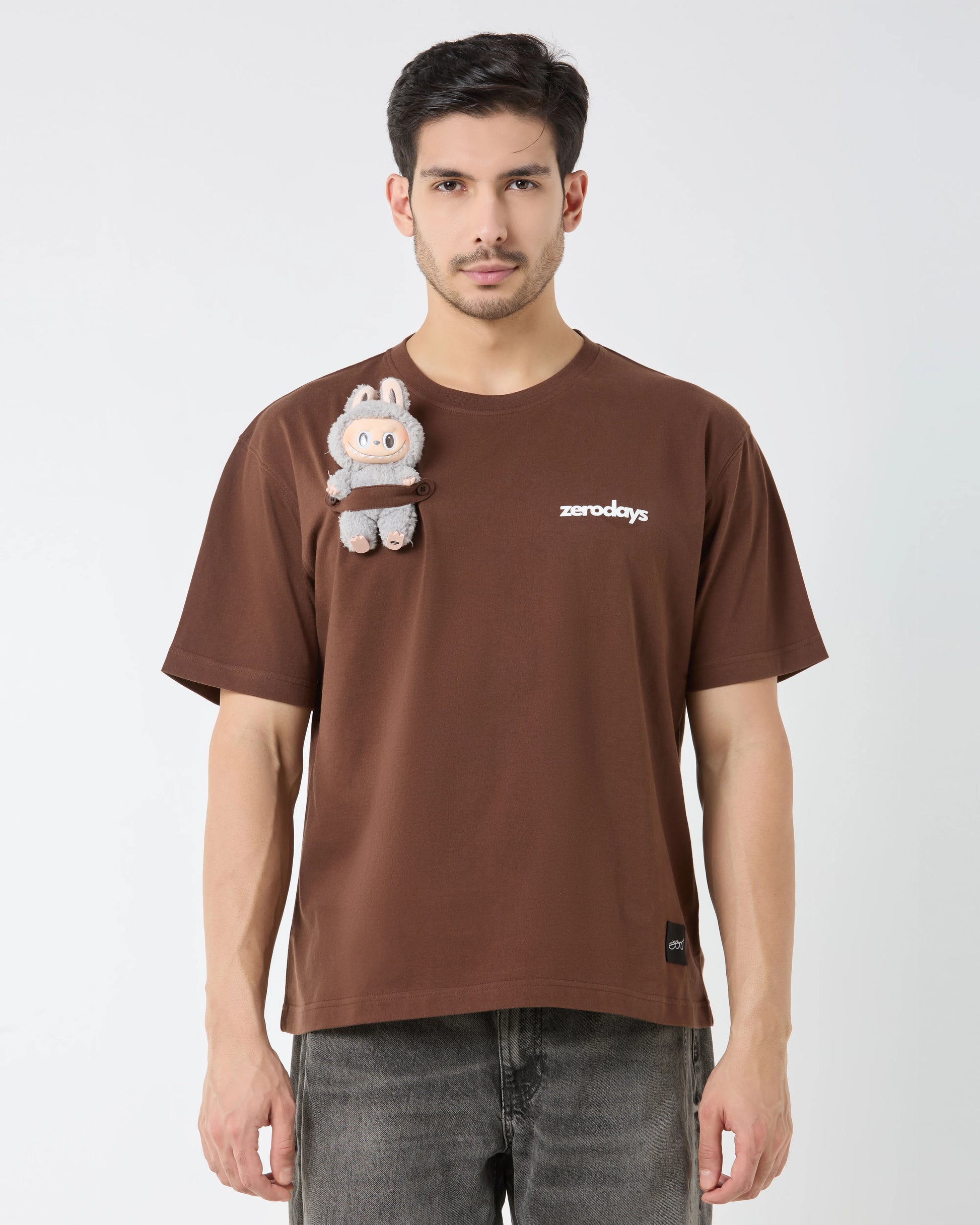 Man wearing a brown t-shirt with a small plush toy attached, on a plain background