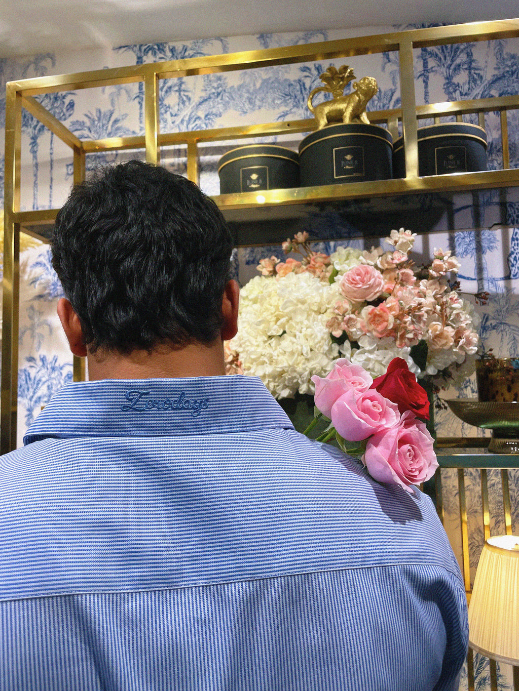 A creatively directed backshot captures an individual wearing the pinstripe shirt, highlighting the Zerodays embroidery on the back of the collar. The aesthetic background complements the scene, while a hint of a bouquet of roses adds a charming touch.
